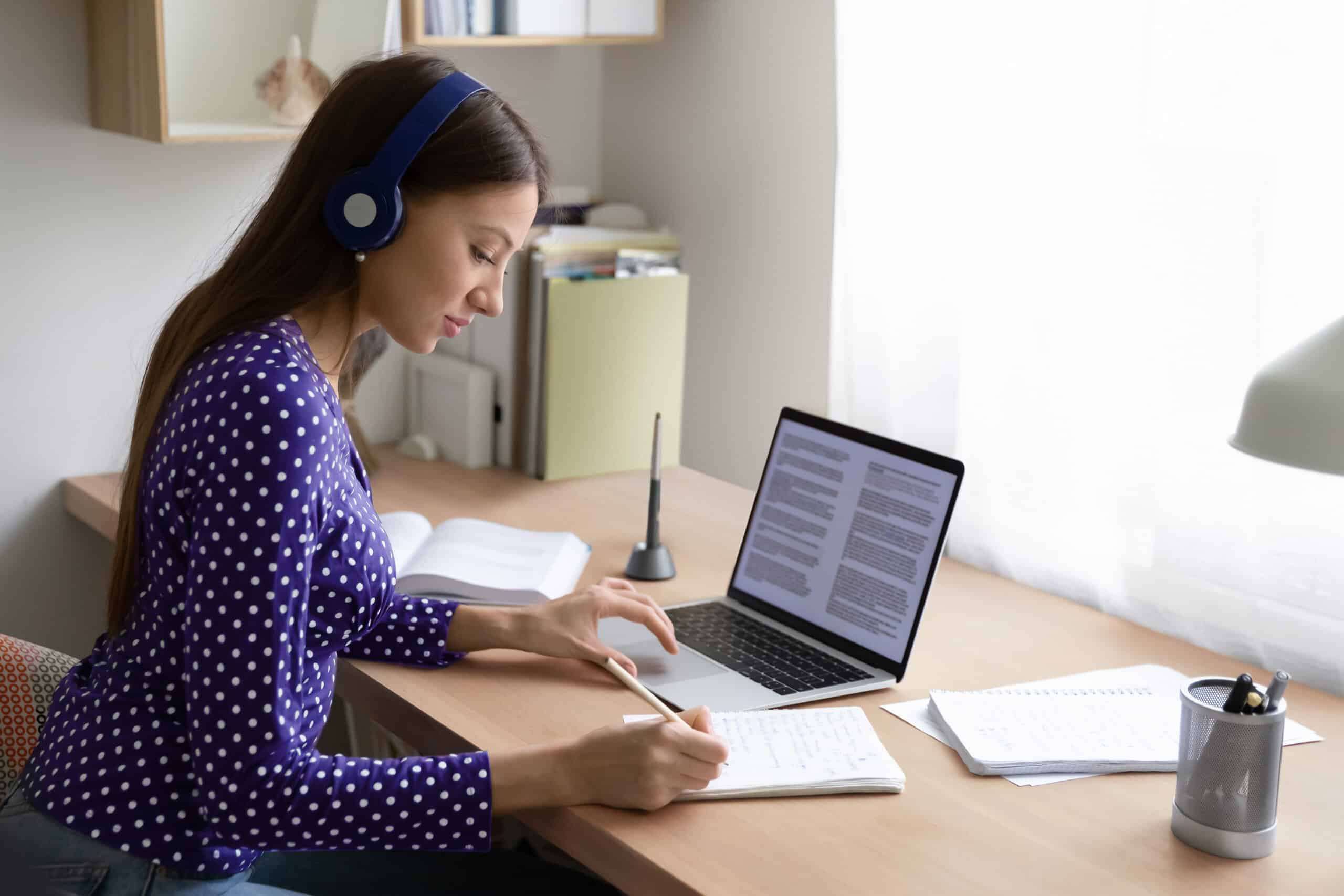 person sitting at desk in front of laptop, writing in notebook
